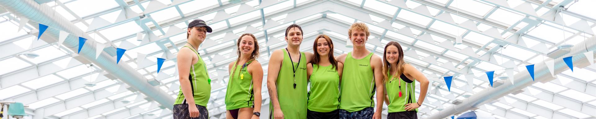 Four lifeguards in uniform sitting on the pool deck facing the water looking back at the camera.
